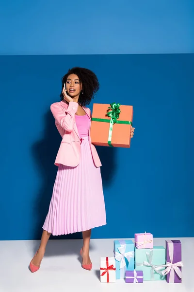 Элегантная улыбающаяся африканская американка разговаривает на смартфоне рядом с подарками на синем фоне — стоковое фото