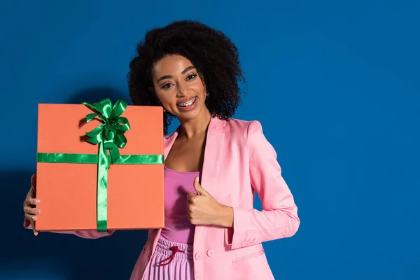 Elegant lächelnde afrikanisch-amerikanische Frau mit Geschenk, das den Daumen nach oben auf blauem Hintergrund zeigt — Stockfoto