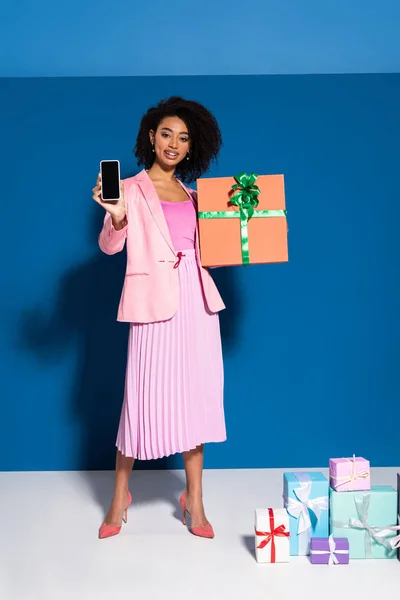 Elegante donna afroamericana sorridente con regalo che mostra smartphone con schermo bianco su sfondo blu — Foto stock