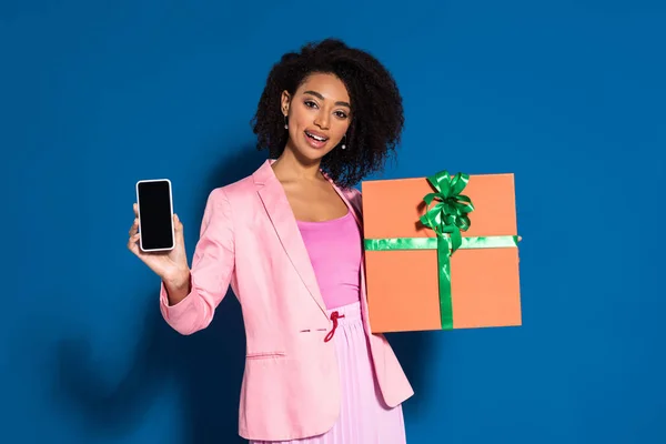 Elegant lächelnde afrikanisch-amerikanische Frau mit Geschenk, das Smartphone mit leerem Bildschirm auf blauem Hintergrund zeigt — Stockfoto