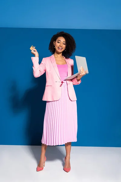 Elegant african american businesswoman holding credit card and laptop on blue background — Stock Photo