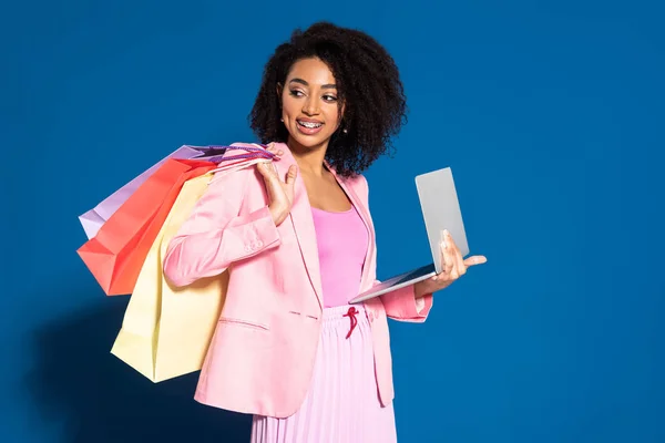 Sonriente elegante mujer de negocios afroamericana con bolsas de compras utilizando el ordenador portátil sobre fondo azul - foto de stock