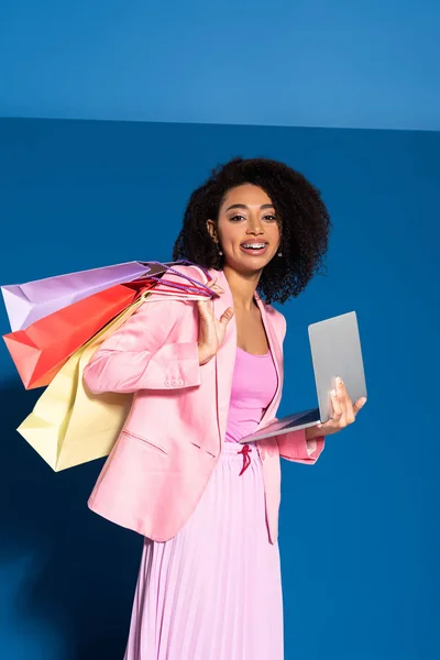 Sonriente elegante mujer de negocios afroamericana con bolsas de compras utilizando el ordenador portátil sobre fondo azul - foto de stock