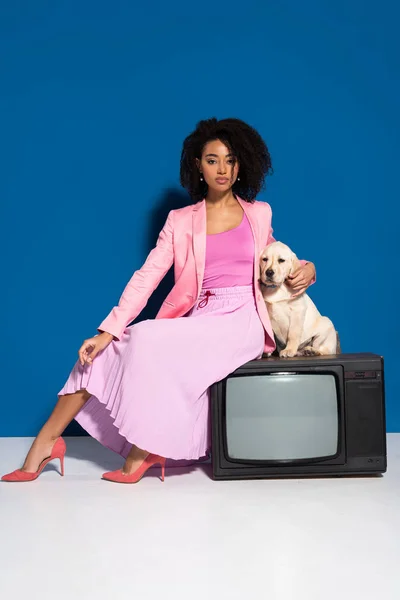 Africano americano mulher sentado no vintage tv com golden retriever filhote de cachorro no fundo azul — Fotografia de Stock