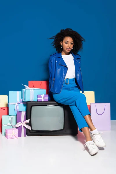 Улыбающаяся африканская американка, сидящая на винтажном телевизоре рядом с подарками и пакетами для покупок на синем фоне — стоковое фото