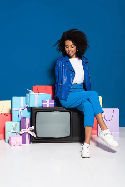 Femme afro-américaine souriante assis sur la télévision vintage près de cadeaux et sacs à provisions sur fond bleu — Photo de stock