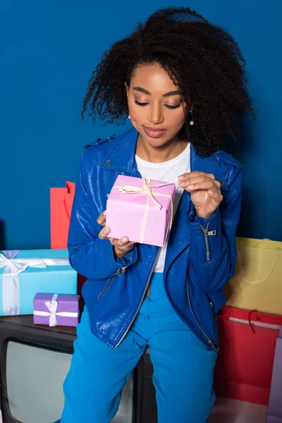 Femme afro-américaine souriante assise sur une télévision vintage avec un cadeau sur fond bleu — Photo de stock