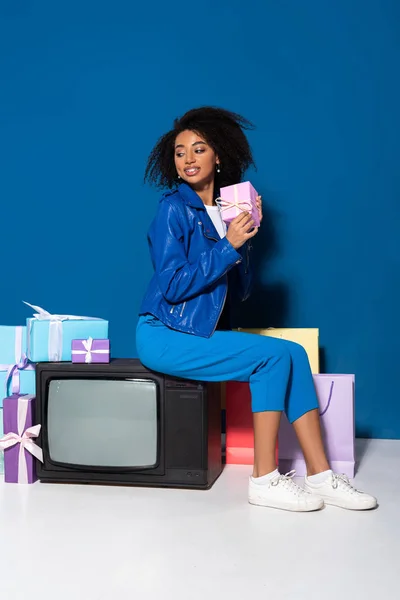Lächelnde afrikanisch-amerikanische Frau sitzt im Vintage-Fernseher neben Geschenken und Einkaufstüten auf blauem Hintergrund — Stockfoto