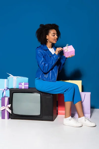 Улыбающаяся африканская американка, сидящая на старинном телевизоре и открывающая подарок на синем фоне — стоковое фото
