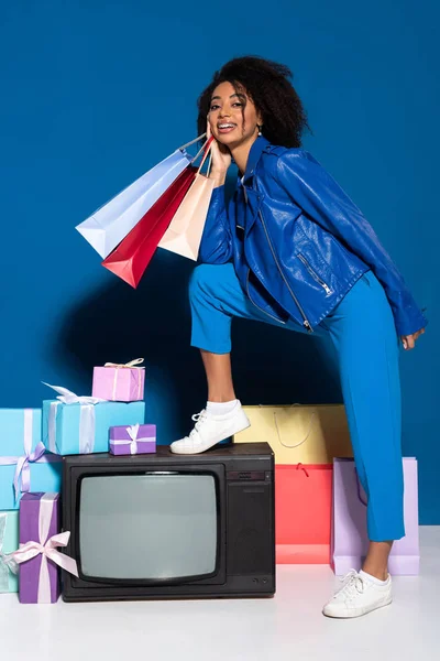 Smiling african american woman sitting on vintage television with shopping bags on blue background — Stock Photo