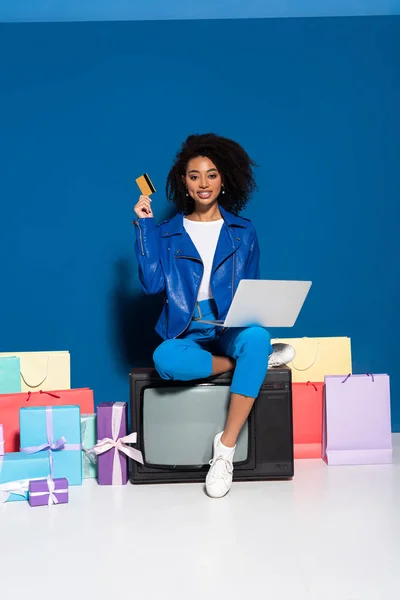 Sonriente mujer afroamericana sentada en la televisión vintage con computadora portátil y tarjeta de crédito cerca de regalos y bolsas de compras sobre fondo azul - foto de stock
