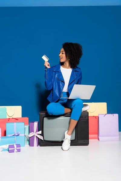 Smiling african american woman sitting on vintage television with laptop and credit card near gifts and shopping bags on blue background — Stock Photo