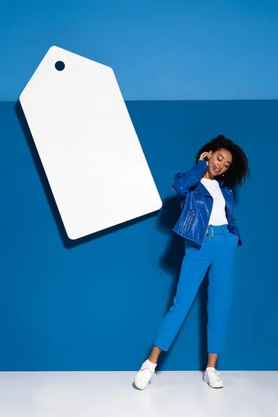 Sorridente donna afro-americana in posa vicino grande prezzo in bianco tag su sfondo blu — Foto stock