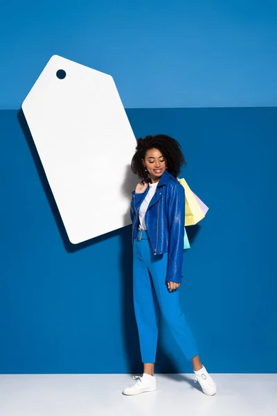 Sorridente mulher americana africana com sacos de compras perto de grande preço em branco no fundo azul — Fotografia de Stock