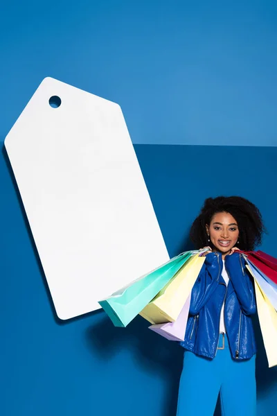 Souriant femme afro-américaine avec des sacs à provisions près de grand prix blanc sur fond bleu — Photo de stock