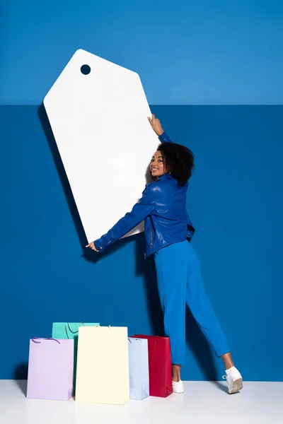 Sonriente mujer afroamericana sosteniendo gran etiqueta de precio en blanco cerca de la bolsa de compras sobre fondo azul - foto de stock