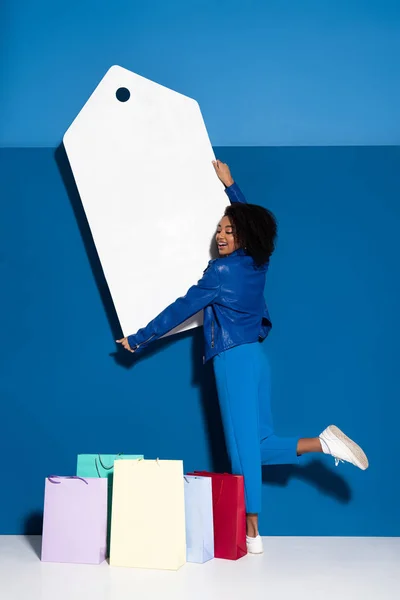 Улыбающаяся африканская американка держит большой пустой ценник рядом с сумкой для покупок на синем фоне — стоковое фото