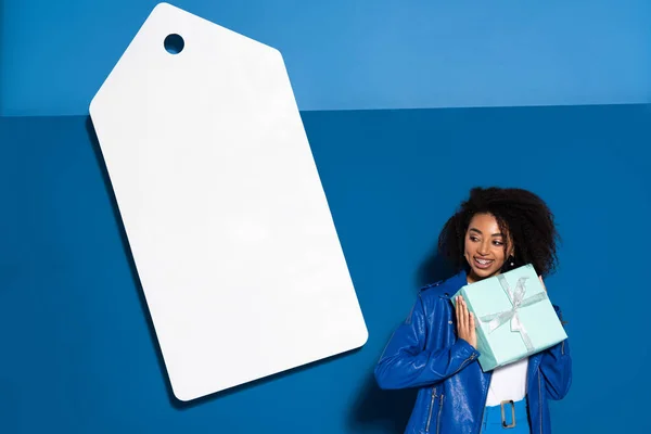 Sorridente mulher americana africana com presente perto de grande preço em branco no fundo azul — Fotografia de Stock