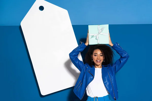 Lächelnde afrikanisch-amerikanische Frau mit Geschenk in der Nähe großer leerer Preisschilder auf blauem Hintergrund — Stockfoto