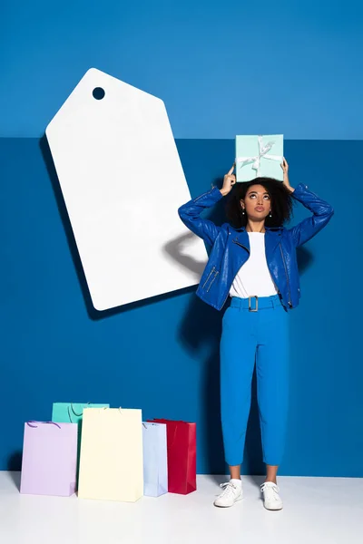Afro-américaine femme avec cadeau près de grand prix blanc étiquette et achats sur fond bleu — Photo de stock