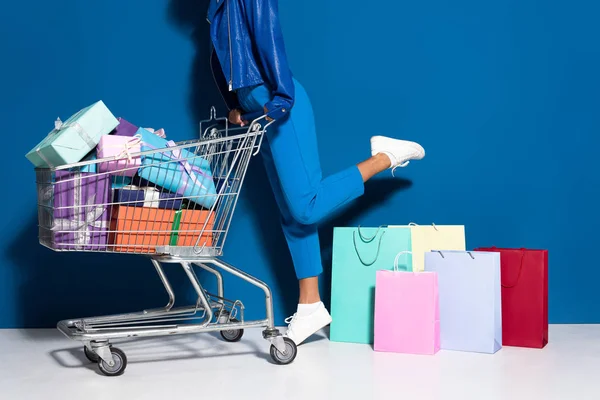Vista recortada de mujer afroamericana con carrito de compras lleno de regalos cerca de bolsas de compras sobre fondo azul - foto de stock