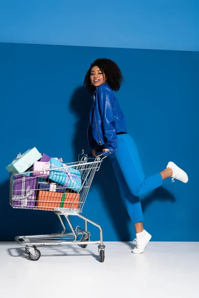 Sonriente mujer afroamericana con carrito de compras lleno de regalos sobre fondo azul - foto de stock