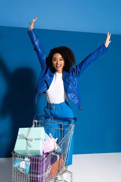 Mujer afroamericana feliz con las manos en el aire y carrito de compras lleno de regalos sobre fondo azul - foto de stock