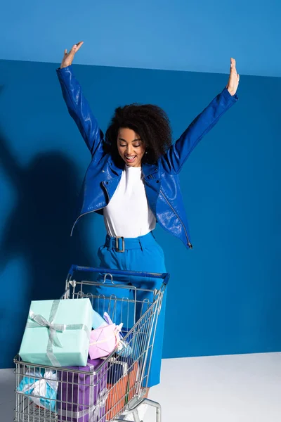 Happy african american woman with hands in air and shopping cart full of gifts on blue background — Stock Photo