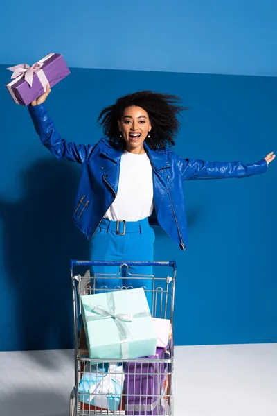 Mujer afroamericana feliz con carrito de compras lleno de regalos sobre fondo azul - foto de stock