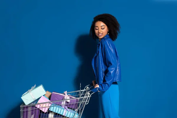Side view of happy african american woman with shopping cart full of gifts on blue background — Stock Photo