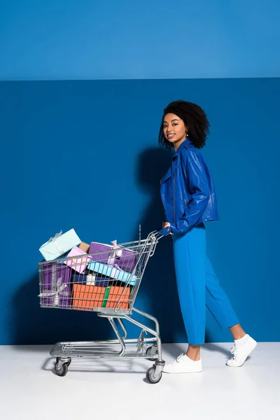 Seitenansicht von glücklichen afrikanisch-amerikanischen Frau mit Einkaufswagen voller Geschenke auf blauem Hintergrund — Stockfoto