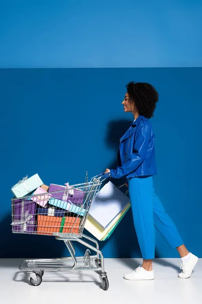 Side view of happy african american woman with shopping cart full of gifts on blue background — Stock Photo