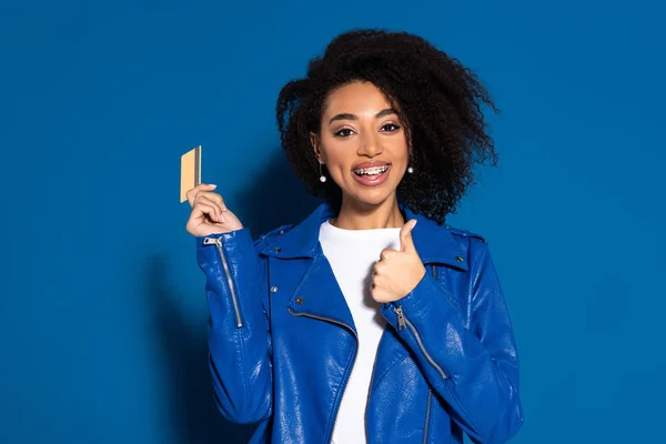 Mujer afroamericana feliz con tarjeta de crédito que muestra el pulgar hacia arriba sobre fondo azul - foto de stock