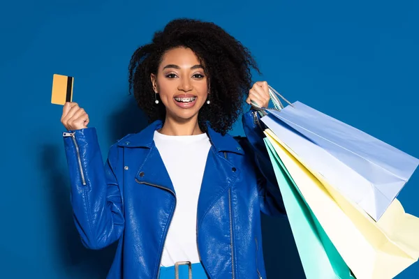 Mulher americana africana feliz com cartão de crédito e sacos de compras em fundo azul — Fotografia de Stock