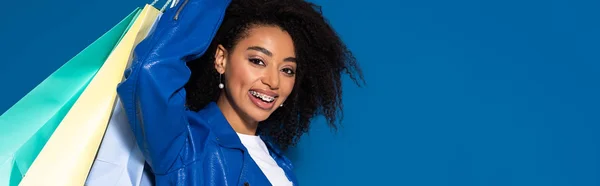 Sonriente mujer afroamericana con bolsas aisladas sobre fondo azul, plano panorámico - foto de stock