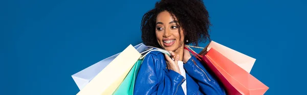 Sonriente mujer afroamericana con bolsas de compras aisladas en azul, tiro panorámico - foto de stock