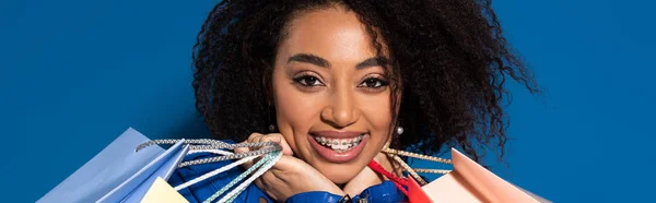 Donna afroamericana sorridente con bretelle dentarie e borse della spesa isolate sul blu, scatto panoramico — Foto stock