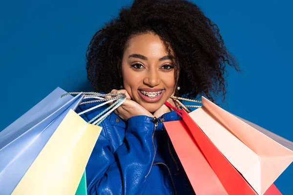Усміхнена афроамериканська жінка з брекетами та сумками ізольована на синьому — стокове фото