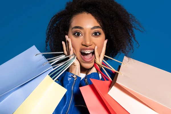 Chocado afro-americano mulher com sacos de compras isolados em azul — Fotografia de Stock