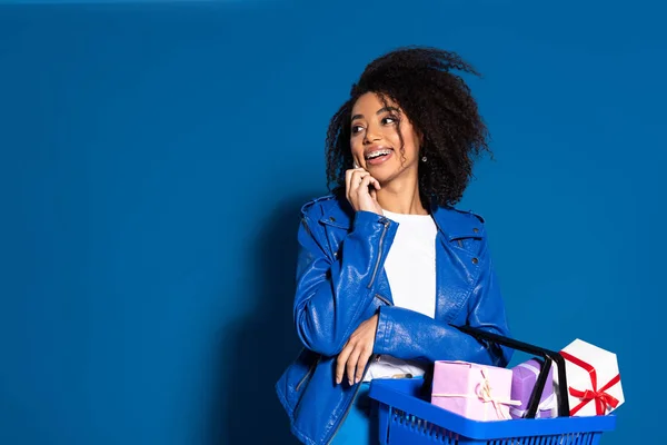 Smiling african american woman holding shopping basket with gifts and talking on smartphone on blue background — Stock Photo
