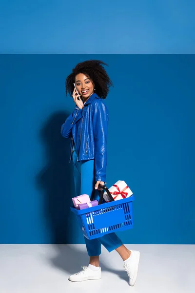 Femme afro-américaine souriante tenant panier avec des cadeaux et parlant sur smartphone sur fond bleu — Photo de stock