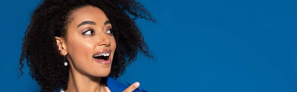 Heureuse femme afro-américaine avec la bouche ouverte pointant du doigt isolé sur bleu, plan panoramique — Photo de stock