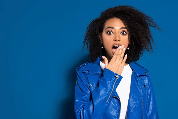 Shocked african american woman coveting mouth with hand on blue background — Stock Photo