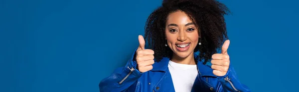 Femme afro-américaine souriante montrant pouces sur fond bleu, vue panoramique — Photo de stock