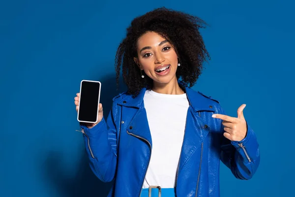 Lächelnde Afroamerikanerin zeigt mit dem Finger auf Smartphone mit leerem Bildschirm auf blauem Hintergrund — Stockfoto