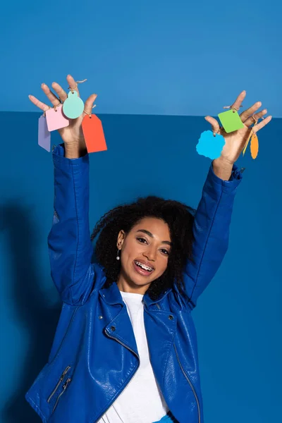 Heureuse femme afro-américaine avec des étiquettes vides sur les mains sur fond bleu — Photo de stock