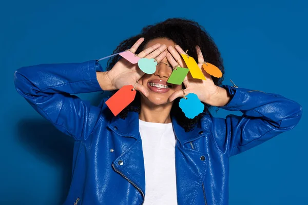 Femme afro-américaine souriante couvrant les yeux avec des étiquettes vides sur les mains sur fond bleu — Photo de stock