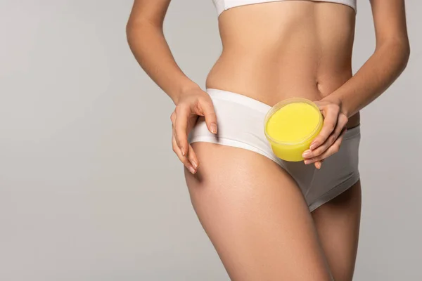 Cropped view of slim girl applying yellow salt scrub from plastic container, isolated on grey — Stock Photo