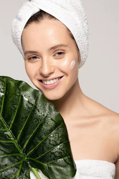 Glückliches Mädchen mit Feuchtigkeitscreme im Gesicht und Handtuch auf dem Kopf posiert mit grünem Blatt, isoliert auf grau — Stockfoto