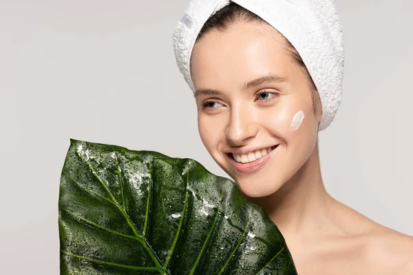 Chica feliz con crema cosmética en la cara y toalla en la cabeza posando con hoja verde, aislado en gris - foto de stock